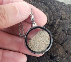 a hand holding a small round pendant with sand inside it on a chain that is attached to a piece of wood