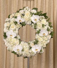 a wreath with white flowers and greenery on a stand in front of a curtain