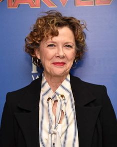 an older woman with curly hair wearing a black jacket and white shirt, standing in front of a blue background