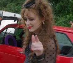 a woman with long red hair standing in front of a car giving the peace sign