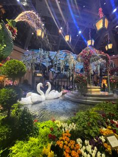 two white swans sitting on top of a fountain surrounded by flowers and greenery in a building