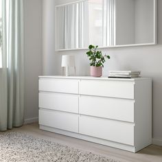 a white dresser with a plant and mirror on it's top, in front of a window