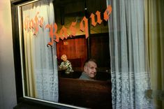 the reflection of a man sitting in front of a window with curtains on both sides