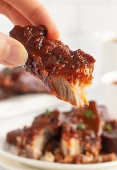 a hand holding up a piece of meat with sauce on it and another plate in the background