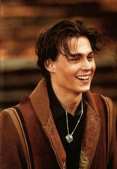 a young man smiling while wearing a brown jacket and black shirt with a necklace on his neck