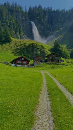 a dirt road going through a lush green field next to a mountain with a waterfall in the background