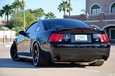 the rear end of a black sports car parked in a parking lot with palm trees