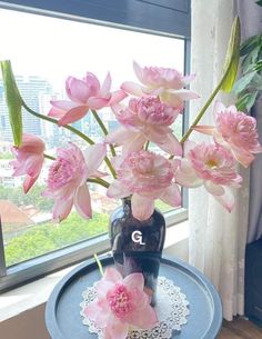 pink flowers are in a vase on a blue tray near a window with lace doily