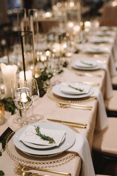 the table is set with white plates and silverware, gold rimmed cutlery, candles and greenery