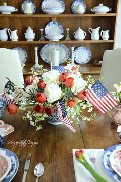 an american flag centerpiece on a dining room table with flowers and patriotic plates in the background