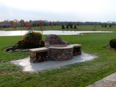 a stone fire pit sitting in the middle of a lush green field next to a pond