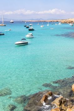 many boats are in the clear blue water near rocks and sand on the shore line