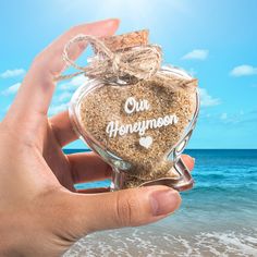 someone is holding up a heart shaped jar with sand in it and the ocean behind them