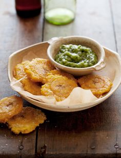 a bowl filled with fried bananas next to a cup of green sauce on top of a wooden table