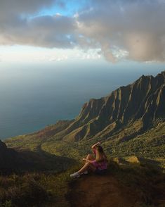 a woman sitting on top of a lush green hillside next to the ocean under a cloudy sky