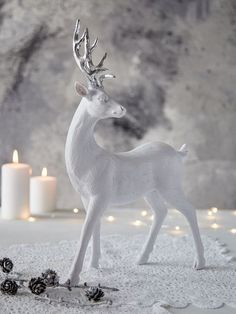 a white deer figurine standing in the snow with pine cones and candles behind it