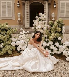 a woman in a white dress sitting on the ground next to some flowers and bushes