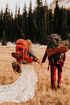 a bride and groom holding hands while walking through the grass with backpacks on their back