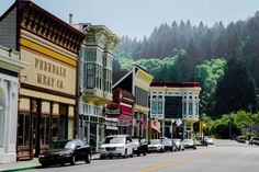 several cars parked on the side of a road in front of buildings and trees with mountains in the background