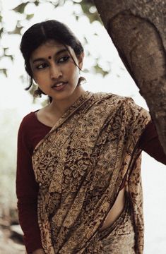 a woman in a sari standing next to a tree trunk and looking at the camera