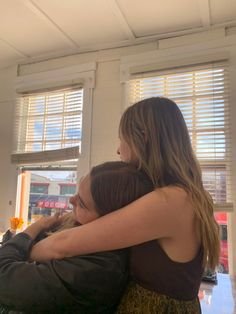 two young women hugging each other in front of windows with blinds on the window sill