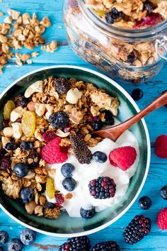 a bowl filled with granola and fruit on top of a blue table next to a jar of yogurt