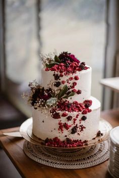 a three tiered white cake with red flowers on top and greenery sprinkled all over it