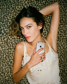 a young woman holding an iphone in her right hand and posing for the camera while wearing a white tank top