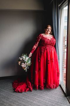 a woman in a red dress standing next to a window and holding a flower bouquet