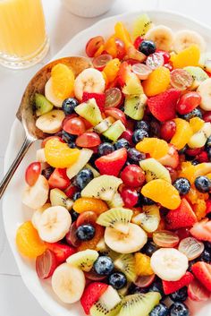 a white plate topped with fruit salad next to a glass of orange juice and a spoon