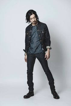 a man with long hair standing in front of a white background wearing black jeans and a denim jacket