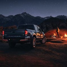 a man standing next to a truck in the desert at night with an open campfire