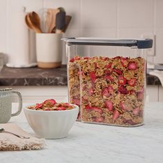 a glass container filled with granola next to a bowl of cereal on a counter