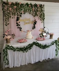 a table topped with cake and desserts next to a pink wall covered in greenery