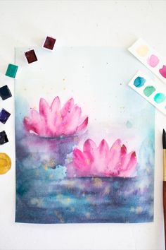 some watercolors and paint sitting on top of a white table with two pink flowers