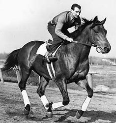 a man riding on the back of a brown horse down a race track in black and white