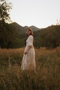 a woman in a white dress is standing in tall grass and looking off into the distance