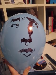 a person holding up a balloon with a drawing on it's face in front of a bookshelf