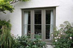 an open window on the side of a white building with green plants and flowers around it