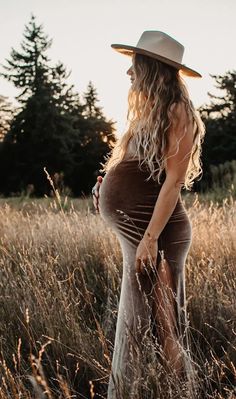 a pregnant woman standing in tall grass wearing a hat