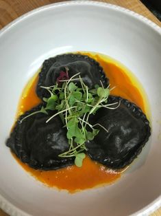 a white bowl filled with black dumplings covered in sauce and garnished with green sprouts