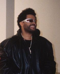 a man with sunglasses on his face standing in front of a wall wearing a leather jacket