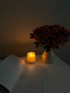 an open book with a lit candle on it next to a vase filled with roses