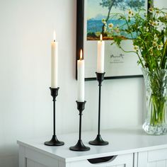 three candles are sitting on a table next to a vase with flowers and a painting