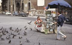 a woman sitting on the ground surrounded by pigeons in front of a man with an umbrella