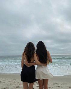 two women are standing on the beach facing the ocean and one has her back to the camera