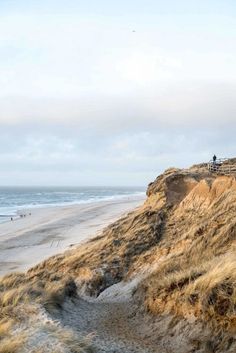 people are walking on the beach near some sand dunes and water in the ocean behind them