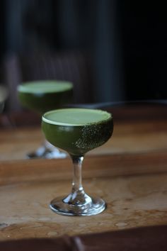 two glasses filled with green liquid sitting on top of a wooden table next to each other