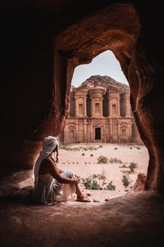 a woman sitting on the ground in front of a rock formation with a building in the background