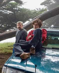 two people sitting on the hood of a blue car in front of a fallen tree